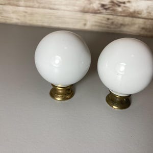 May include: Two white, spherical cabinet knobs with a glossy finish. Each knob is mounted on a brass-colored base. The knobs are likely for furniture or decorative purposes.