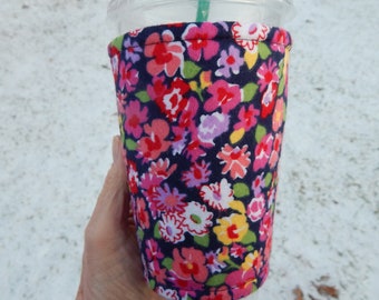 May include: A reusable cup cozy with a floral print in shades of pink, red, yellow, and white on a navy blue background. The cozy is made of fabric and has a sewn seam around the top edge.