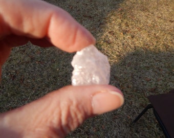 May include: A clear, translucent, pink crystal with a rough, irregular shape. The crystal is being held in a hand.