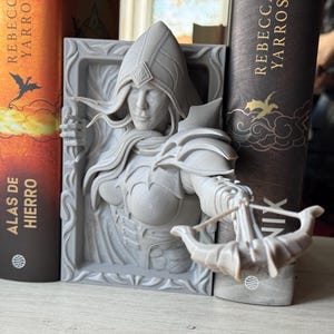 May include: A grey 3D printed bookend featuring a female figure with a hood and a crossbow. The figure is holding the crossbow in her right hand and is looking to the right. The bookend is set against a background of two books.