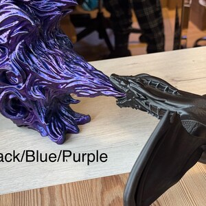May include: A black dragon figurine with wings outstretched, breathing purple fire. The fire is a separate figurine with a blue and purple iridescent finish. The text "Black/Blue/Purple" is visible in the image.