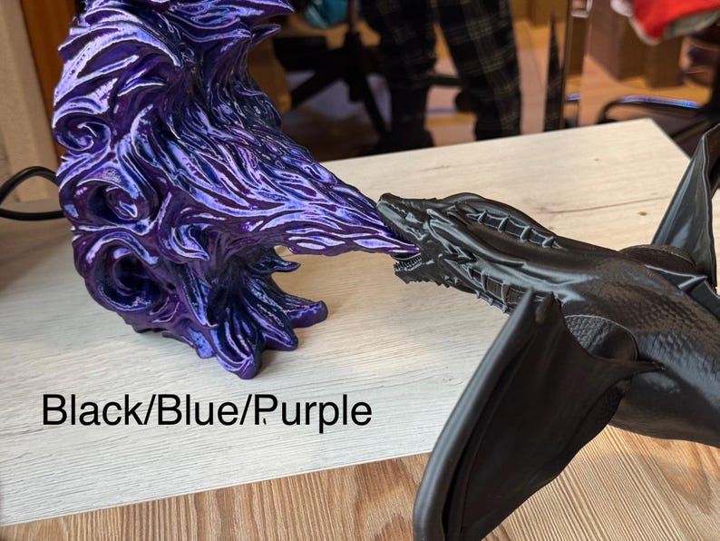 May include: A black dragon figurine with wings outstretched, breathing purple fire. The fire is a separate figurine with a blue and purple iridescent finish. The text "Black/Blue/Purple" is visible in the image.