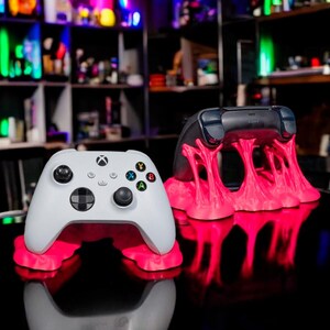 Könnte beinhalten: Zwei weiße Videospiel-Controller auf rosa, 3D-gedruckten Ständern. Die Controller befinden sich auf einer schwarzen Oberfläche mit einer Reflexion der Controller und Ständer.
