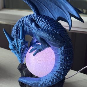 Puede incluir: Una figura de dragón azul con alas extendidas, posada sobre una base rocosa negra. El dragón sostiene una luna esférica y brillante. La escultura está iluminada con una suave luz púrpura, creando una pieza decorativa de temática fantástica.