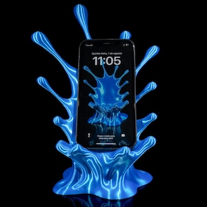 以下が含まれることがあります： 水しぶきのような形をした青い3Dプリントのスマートフォンスタンド。スタンドは、青い水しぶきの画面と画面に表示された時刻11:05のスマートフォンを保持しています。