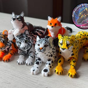May include: A collection of six 3D printed animal figurines. The figurines are a cheetah, a snow leopard, a tiger, a fox, and two white tigers. The figurines are all posed in a sitting or lying down position. The figurines are all made of a white, black, and orange plastic.