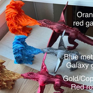May include: Four 3D printed dragon figurines with flames coming out of their mouths. The dragons are painted in different colors: orange/red, blue metallic/galaxy grey, gold/copper/red rose, and gold. The text "Orange/red galaxy", "Blue metallic/Galaxy grey", "Gold/Copper/Red rose" is written below each dragon.