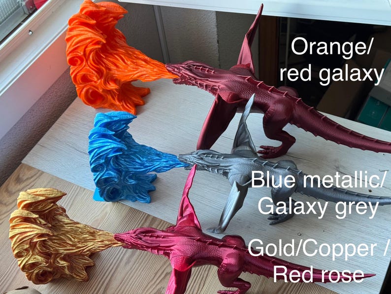 May include: Four 3D printed dragon figurines with flames coming out of their mouths. The dragons are painted in different colors: orange/red, blue metallic/galaxy grey, gold/copper/red rose, and gold. The text "Orange/red galaxy", "Blue metallic/Galaxy grey", "Gold/Copper/Red rose" is written below each dragon.