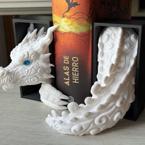 May include: White dragon bookends with blue eyes and detailed features. The dragon head and tail flank a book titled "ALAS DE HIERRO". The bookends are set in black rectangular holders.