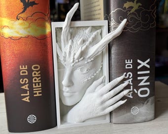 Könnte beinhalten: Weiße, 3D-gedruckte Buchstützen mit einer drachenartigen Kreatur mit Hörnern und Schuppen. Das Gesicht der Kreatur ist teilweise sichtbar, und eine Hand ragt aus dem Rahmen. Die Buchstützen sind auf zwei Büchern mit den Titeln "Alas de Hierro" und "Alas de Ónix" platziert.