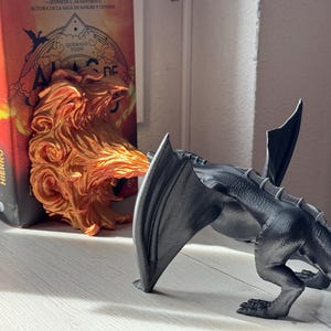 May include: A black dragon bookend with a fire-themed bookend in the background. The bookend is made of plastic and has a detailed design. The bookend is holding up two books with the titles "Alas de Sangre" and "Alas de Hierro".