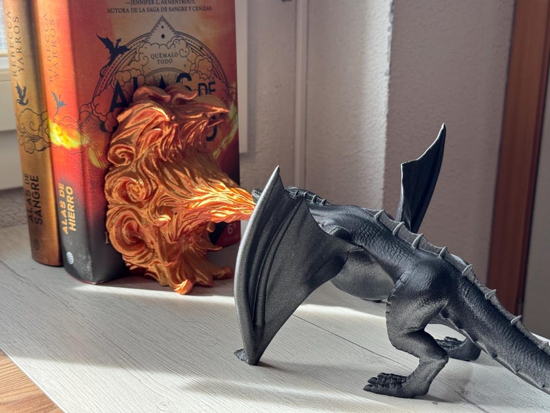 May include: A black dragon bookend with a fire-themed bookend in the background. The bookend is made of plastic and has a detailed design. The bookend is holding up two books with the titles "Alas de Sangre" and "Alas de Hierro".