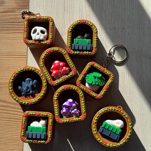 May include: A selection of miniature keyrings with quirky designs. Each keyring has a brown frame with a decorative border and contains a different object, such as a skull, mushrooms, and a cat. The keyrings are displayed on a light-coloured surface.