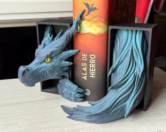 May include: A blue dragon bookend set. The dragon head and tail are detailed with textured scales and yellow eyes. The bookends are black and hold a book with the title "ALAS DE HIERRO".