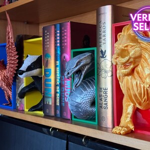 Può includere: Un set di ferma libri con un leone dorato, un serpente nero e argento, un tasso e una fenice. I ferma libri sono esposti su uno scaffale di legno con una varietà di libri, tra cui "Alas de Sangre" di Rebecca Yarros, "Dune" di Frank Herbert e "Hijos de Dune" di Frank Herbert.