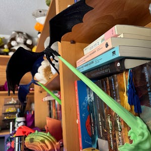 May include: A black dragon figurine with green wings, perched on a bookshelf with a stack of books. The books have titles in Spanish and English, including "Game of Thrones" and "A Clash of Kings".