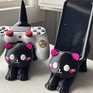 Può includere: Due supporti per telefono e controller a forma di gatto nero. Il supporto per telefono contiene uno smartphone nero. Il supporto per controller contiene un controller PlayStation 5 bianco con pulsanti rossi. Entrambi i supporti hanno orecchie rosa e occhi bianchi.