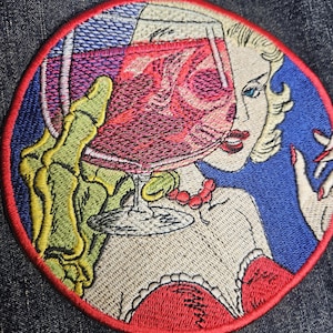 May include: Embroidered patch featuring a retro-style illustration of a woman holding a cocktail. The woman has blonde hair, red lipstick, and a red necklace. A skeleton hand holds the glass. The patch has a red border.