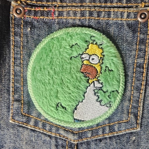 May include: A green and white embroidered patch of a cartoon character with yellow hair and a beard. The character is on a green background with a white outline.
