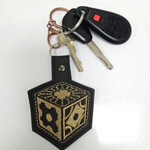 May include: Black leather keychain with a gold embroidered image of a cube with a gear and other geometric designs. The keychain is attached to a set of keys and a key fob.