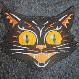 May include: Black embroidered iron-on patch of a cat with yellow eyes, orange and white details, and a toothy grin.