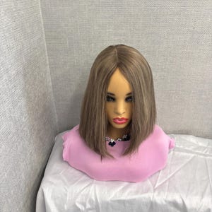 May include: A mannequin head with a light brown wig, styled in a shoulder-length bob. The mannequin is wearing a pink top and a decorative necklace. The background is a light grey wall and a white surface.