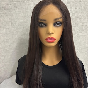 May include: A mannequin head with a dark brown, straight-hair wig. The wig has a lace front and is styled with a centre part. The mannequin is wearing a black t-shirt.