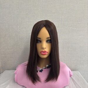 May include: A mannequin head with a brown wig. The wig is styled straight and falls to the shoulders. The mannequin is wearing a pink top and a necklace with dark blue and silver beads. The background is a light grey.
