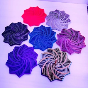 May include: A collection of seven colorful, star-shaped decorative objects. The objects feature a layered, spiral design and are in shades of red, black, blue, purple, silver, and gold. The objects are arranged on a white surface.
