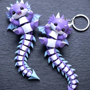 May include: Two purple dragon keychains with white accents and light blue wings and tail fins. The dragons have a happy expression and are attached to a silver keyring. The keychains are made of a smooth material.