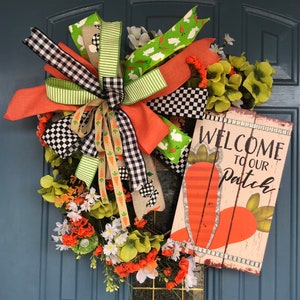 May include: A decorative Easter wreath with a wooden sign that reads "Welcome to our Patch". The wreath features orange, green, and white flowers, and a large bow with various patterned ribbons, including carrots and bunnies.