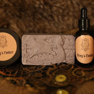 May include: A collection of grooming products with a Viking theme. Includes a round container, a bar of soap with a Viking ship carving, and a dropper bottle. All items have the text "Frey's Ember" on them.