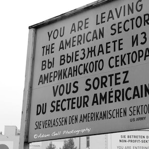 Può includere: Foto in bianco e nero di un cartello con testo in inglese, francese, tedesco e russo. Il cartello recita "You are leaving the American Sector" in inglese, "Vous sortez du secteur Americain" in francese, "Sie verlassen den Amerikanischen Sektor" in tedesco e "Вы выезжаете из американского сектора" in russo. Il cartello è stato affisso dall'esercito americano.