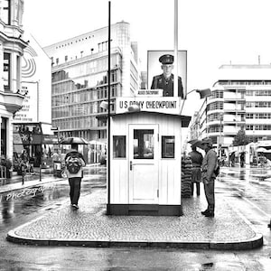 Könnte beinhalten: Ein Schwarzweißfoto eines US-Army-Checkpoints in Berlin, Deutschland. Der Checkpoint ist ein kleines Gebäude mit einem Schild, auf dem "US Army Checkpoint" steht. Menschen gehen am Checkpoint vorbei, einige von ihnen tragen Regenschirme. Das Foto wurde bei Regen aufgenommen.