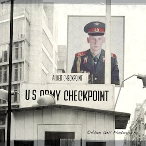 Può includere: Una fotografia in bianco e nero di un posto di blocco a Berlino, Germania. Il cartello recita "US Army Checkpoint". Un soldato in uniforme militare è raffigurato su un cartello sopra il posto di blocco. Il cartello sopra il soldato recita "Allied Checkpoint".