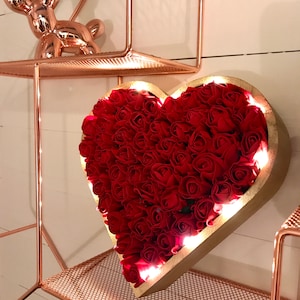 Pode incluir: Um quadro de madeira em forma de cora&ccedil;&atilde;o cheio de rosas de tecido vermelho e iluminado com luzes brancas. O quadro est&aacute; montado em uma prateleira de arame de cobre.