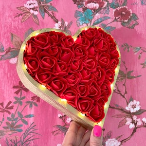 Pode incluir: Um quadro de madeira em forma de cora&ccedil;&atilde;o cheio de rosas de espuma vermelha e contornado com tinta dourada. O quadro tem uma corda de luzes brancas quentes ao redor da borda.