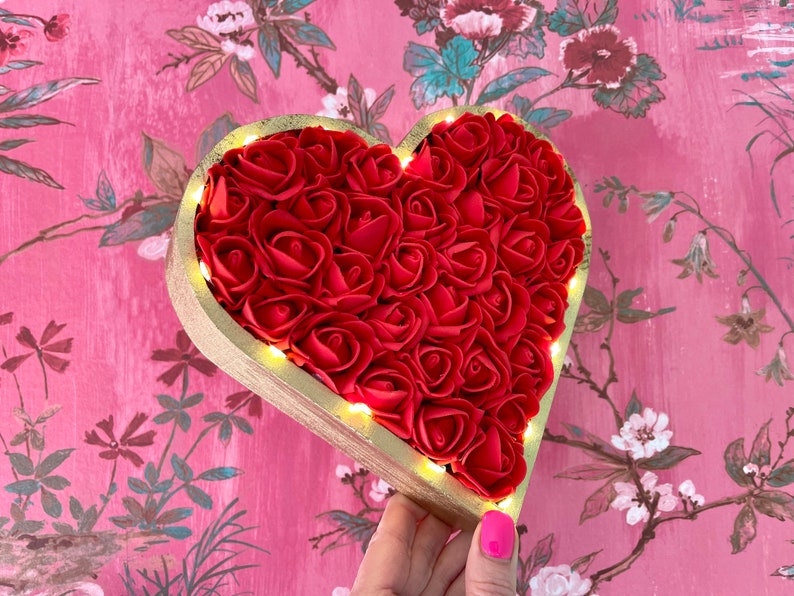 Pode incluir: Um quadro de madeira em forma de cora&ccedil;&atilde;o cheio de rosas de espuma vermelha e contornado com tinta dourada. O quadro tem uma corda de luzes brancas quentes ao redor da borda.