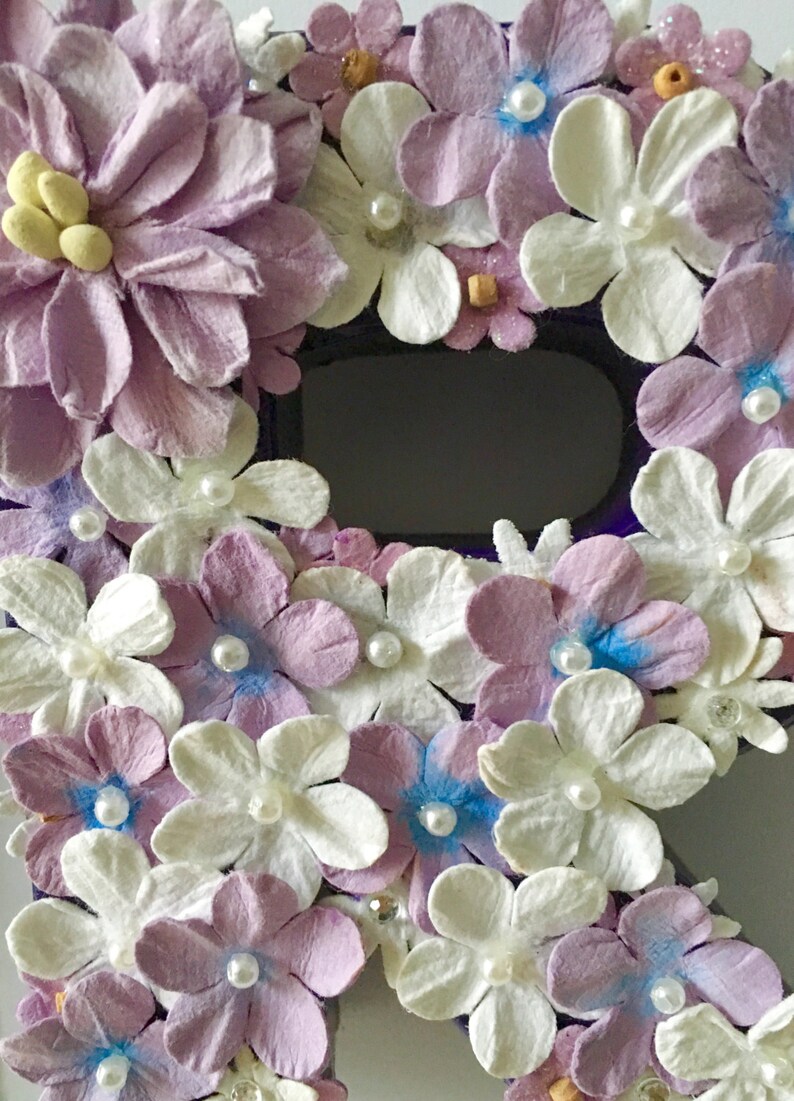 May include: A purple letter R decorated with white and lavender paper flowers. The flowers have pearl accents and some have a light blue center.