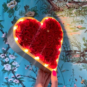 Pode incluir: Um quadro de madeira em forma de cora&ccedil;&atilde;o cheio de rosas artificiais vermelhas e contornado com luzes LED brancas quentes. O quadro &eacute; segurado na m&atilde;o contra um fundo de padr&atilde;o floral azul e verde.