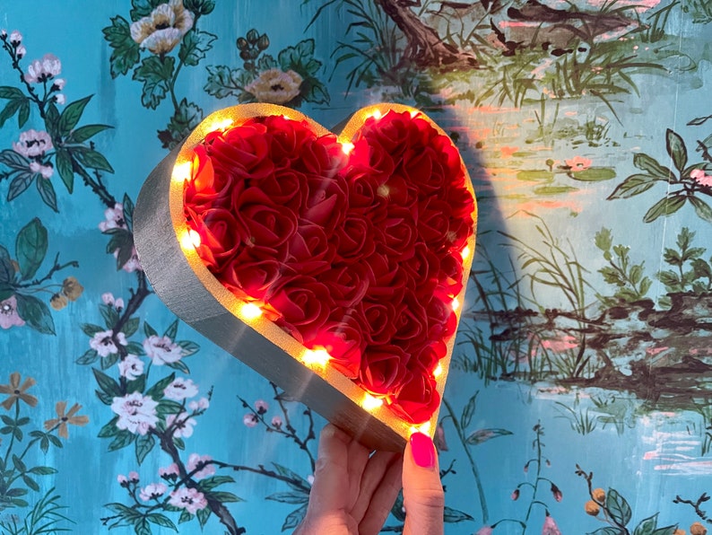 Pode incluir: Um quadro de madeira em forma de cora&ccedil;&atilde;o cheio de rosas artificiais vermelhas e contornado com luzes LED brancas quentes. O quadro &eacute; segurado na m&atilde;o contra um fundo de padr&atilde;o floral azul e verde.