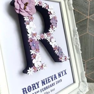 May include: A white frame with a navy blue letter R decorated with purple and white flowers. The text "Rory Nieva Nyx 22nd February 2019 7lbs 6oz" is printed below the letter.