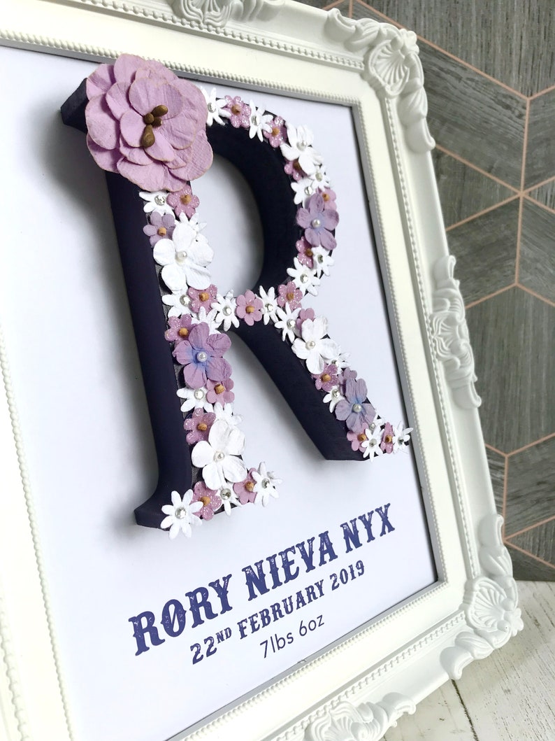 May include: A white frame with a navy blue letter R decorated with purple and white flowers. The text "Rory Nieva Nyx 22nd February 2019 7lbs 6oz" is printed below the letter.