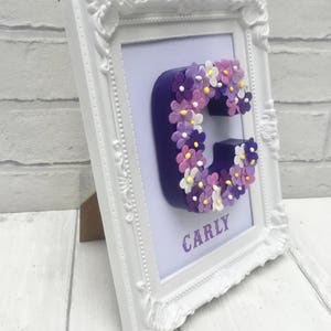 May include: A white frame with a purple letter C decorated with purple, pink, and white flowers. The name "CARLY" is written below the letter.
