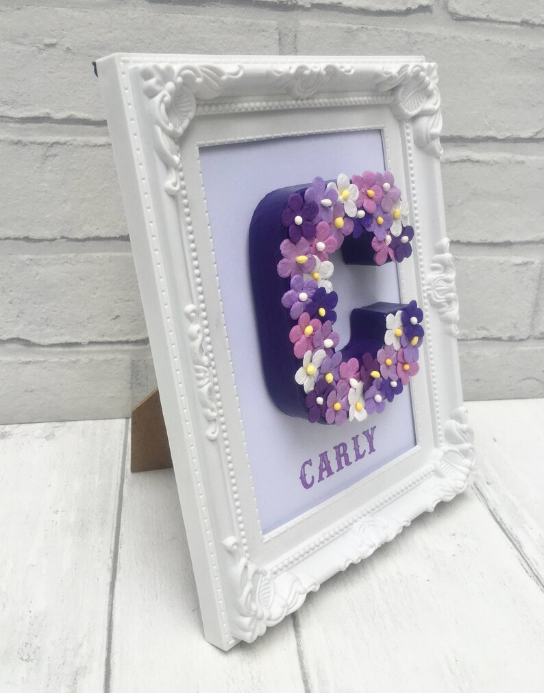 May include: A white frame with a purple letter C decorated with purple, pink, and white flowers. The name "CARLY" is written below the letter.