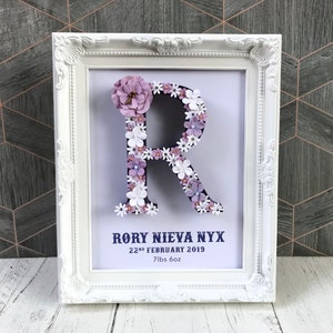 May include: White frame with a white letter R made of purple and white flowers. The frame has the text "Rory Nieva Nyx 22nd February 2019 7lbs 6oz"