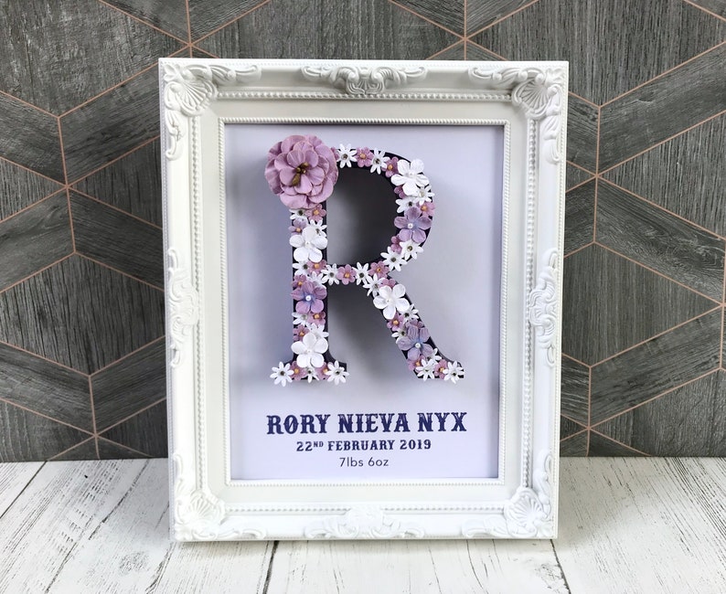 May include: White frame with a white letter R made of purple and white flowers. The frame has the text "Rory Nieva Nyx 22nd February 2019 7lbs 6oz"