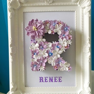 May include: A white frame with a purple letter R made of flowers. The name "RENEE" is written in purple below the letter.