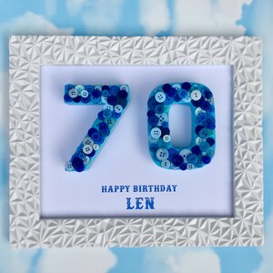 May include: A white frame with a blue and white button art piece that says "70" in the centre. The frame has a geometric pattern. Below the numbers, it says "Happy Birthday Len".
