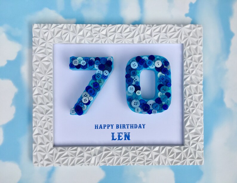 May include: A white frame with a blue and white button art piece that says "70" in the centre. The frame has a geometric pattern. Below the numbers, it says "Happy Birthday Len".